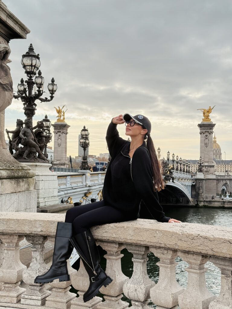 Woman in a black outfit and cap sitting by Pont Alexandre III